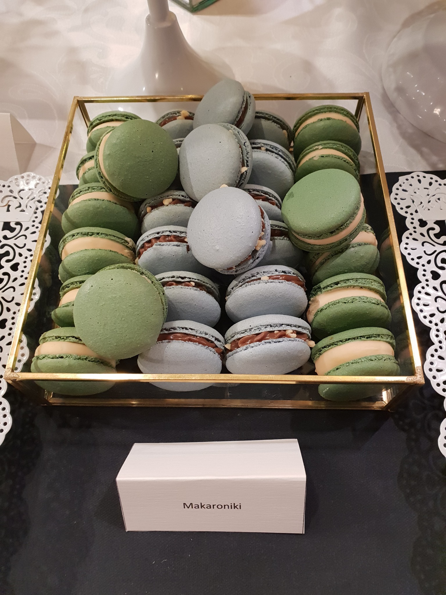 Prezentacja makaroników w odcieniach zieleni i błękitu, ułożonych w złotej, geometrycznej ramce na ciemnym tle, z widoczną etykietą z napisem 'Makaroniki'.