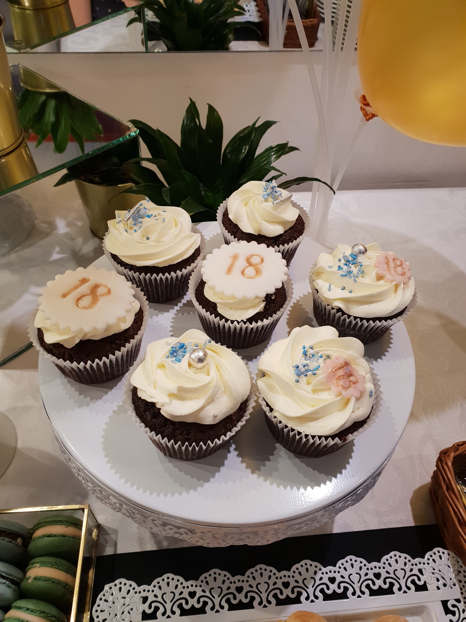 Siedem czekoladowych babeczek z białym kremem i dekoracjami na białym paterze, niektóre z liczbą 18, obok dekoracje urodzinowe i pudełko z makaronikami.