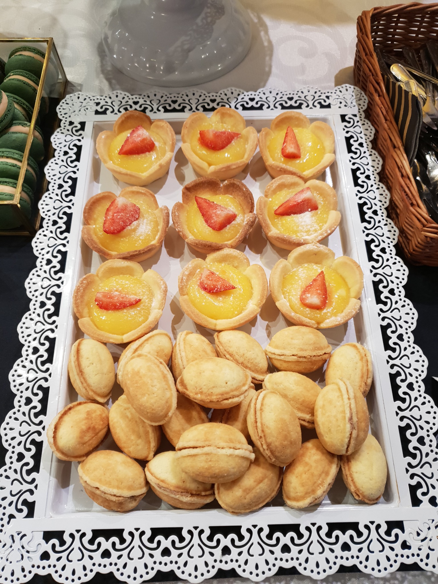 Biała taca z dekoracyjnymi brzegami prezentuje rzędy tartaletek z kremem cytrynowym i truskawkami, obok ułożone ciasteczka orzeszki; w tle widoczne makaroniki i koszyk ze sztućcami.