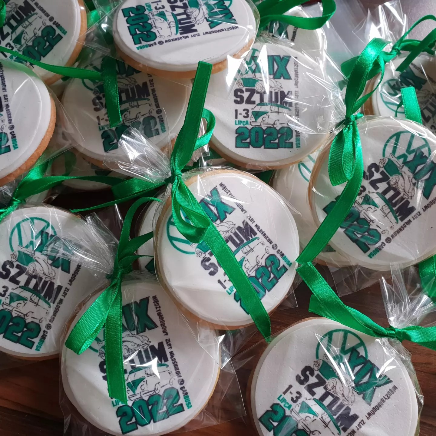 Kolekcja okrągłych ciasteczek zapakowanych w folię, ozdobionych zielonymi wstążkami i nadrukiem z logo zlotu miłośników Garbusa w Sztumie, z datą 1-3 lipca 2022.