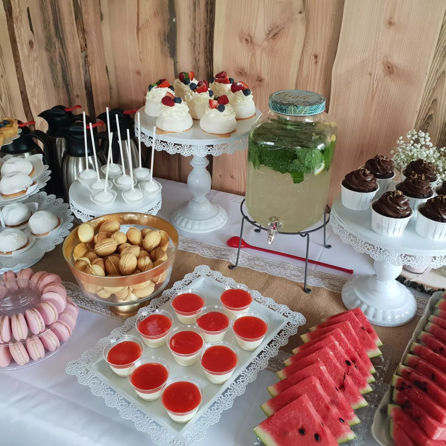 Stół z różnorodnymi deserami: mini bezy z owocami, babeczki czekoladowe, panna cotta z sosem owocowym, arbuz, makaroniki, ciastka orzechy i lemoniada w słoju, ustawione na białym obrusie...