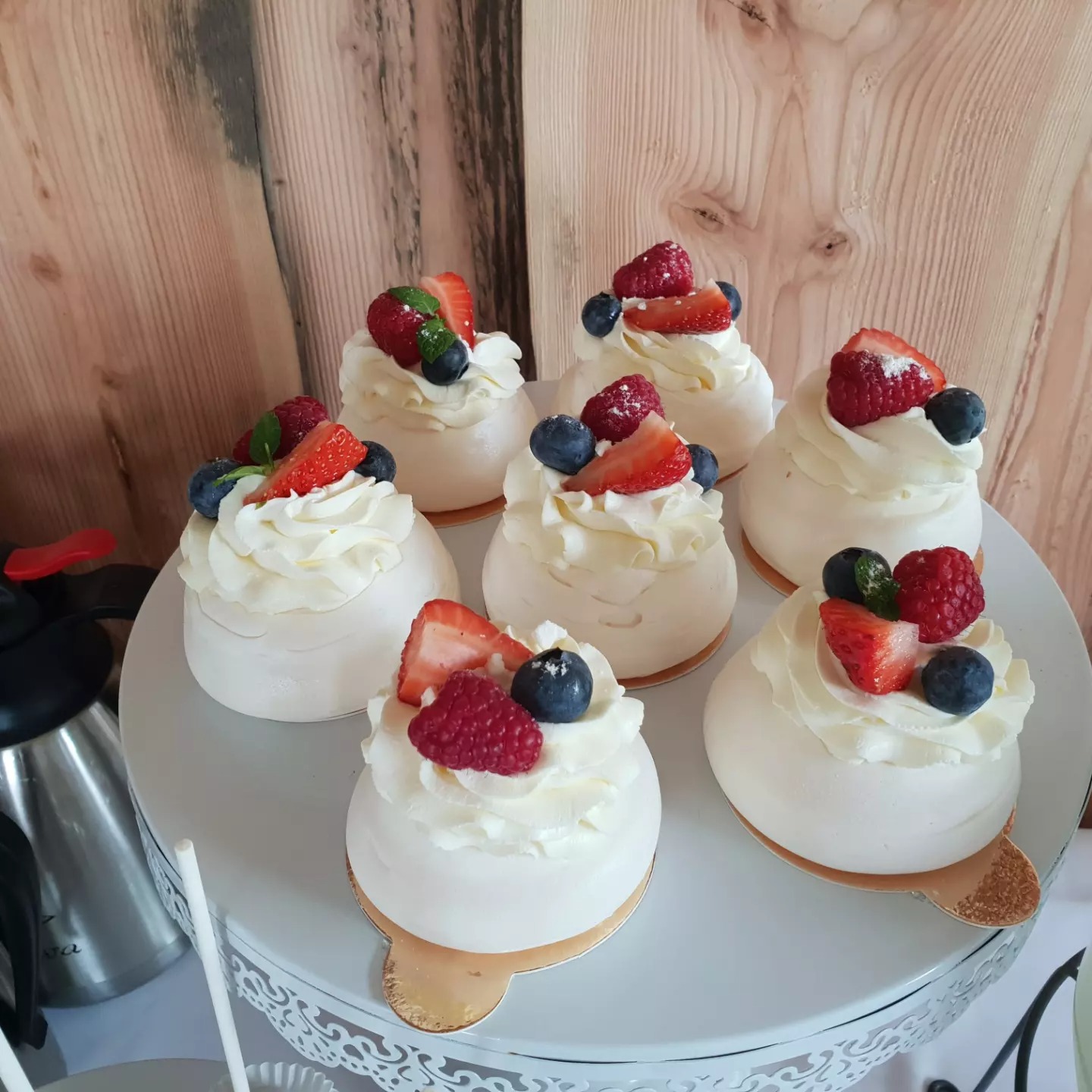 Sześć mini bez z kremem i owocami (truskawki, maliny, borówki) na białym, ażurowym stojaku, drewniane tło.