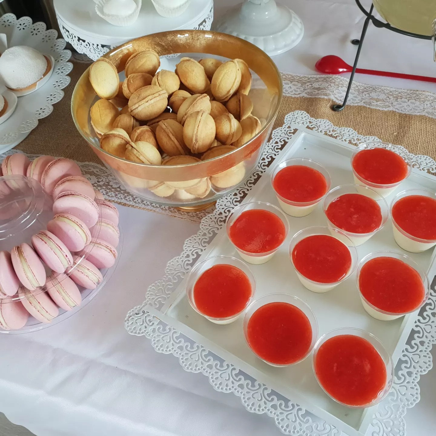 Słodki stół z różowymi makaronikami w przezroczystym pudełku, orzechami w złotej misie i deserami z czerwoną polewą na białym obrusie z koronkową serwetą.