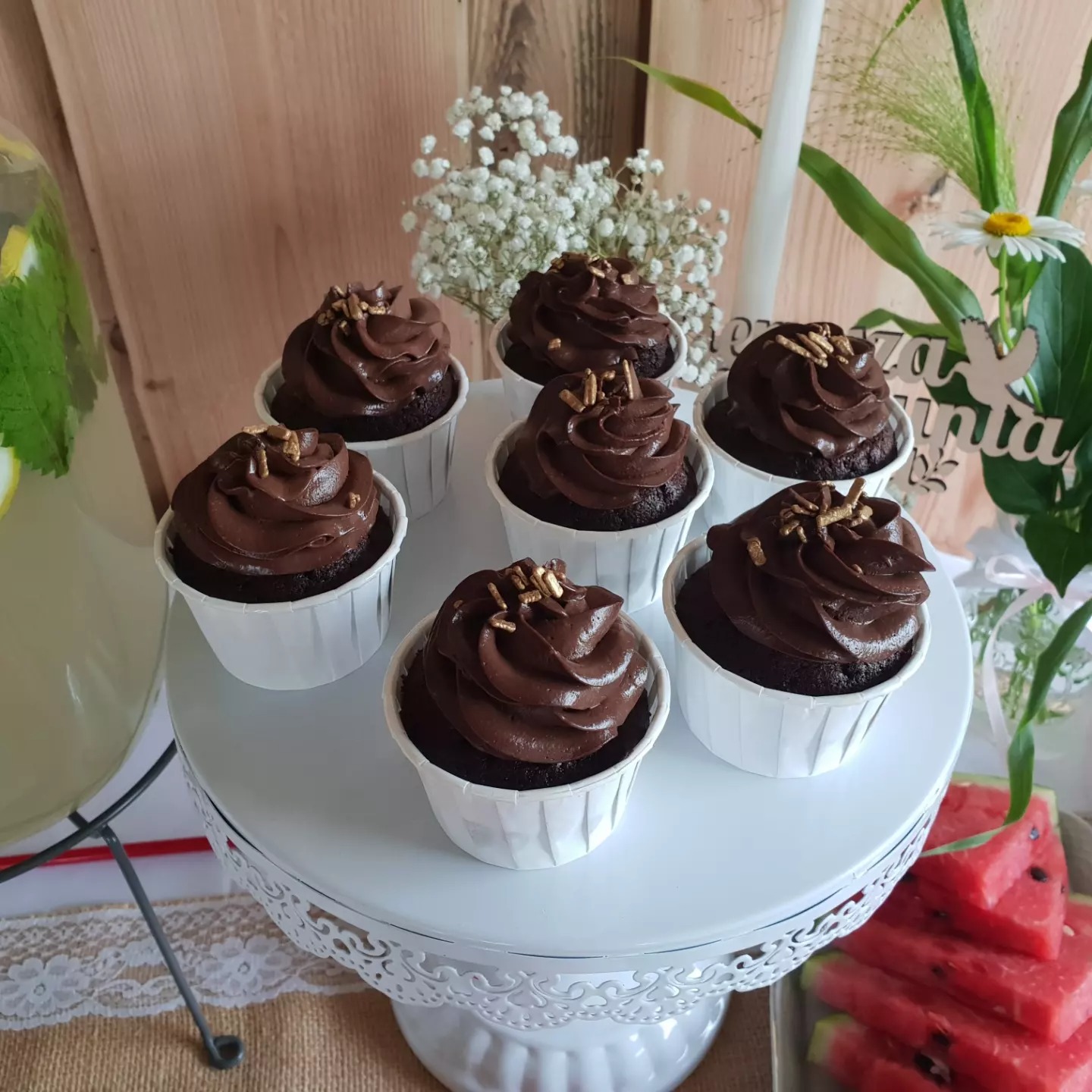 Siedem czekoladowych babeczek z posypką z płatków złota, ustawionych na białym, ażurowym stojaku, obok lemoniady z cytryną i arbuzem, dekoracja kwiatowa w tle.