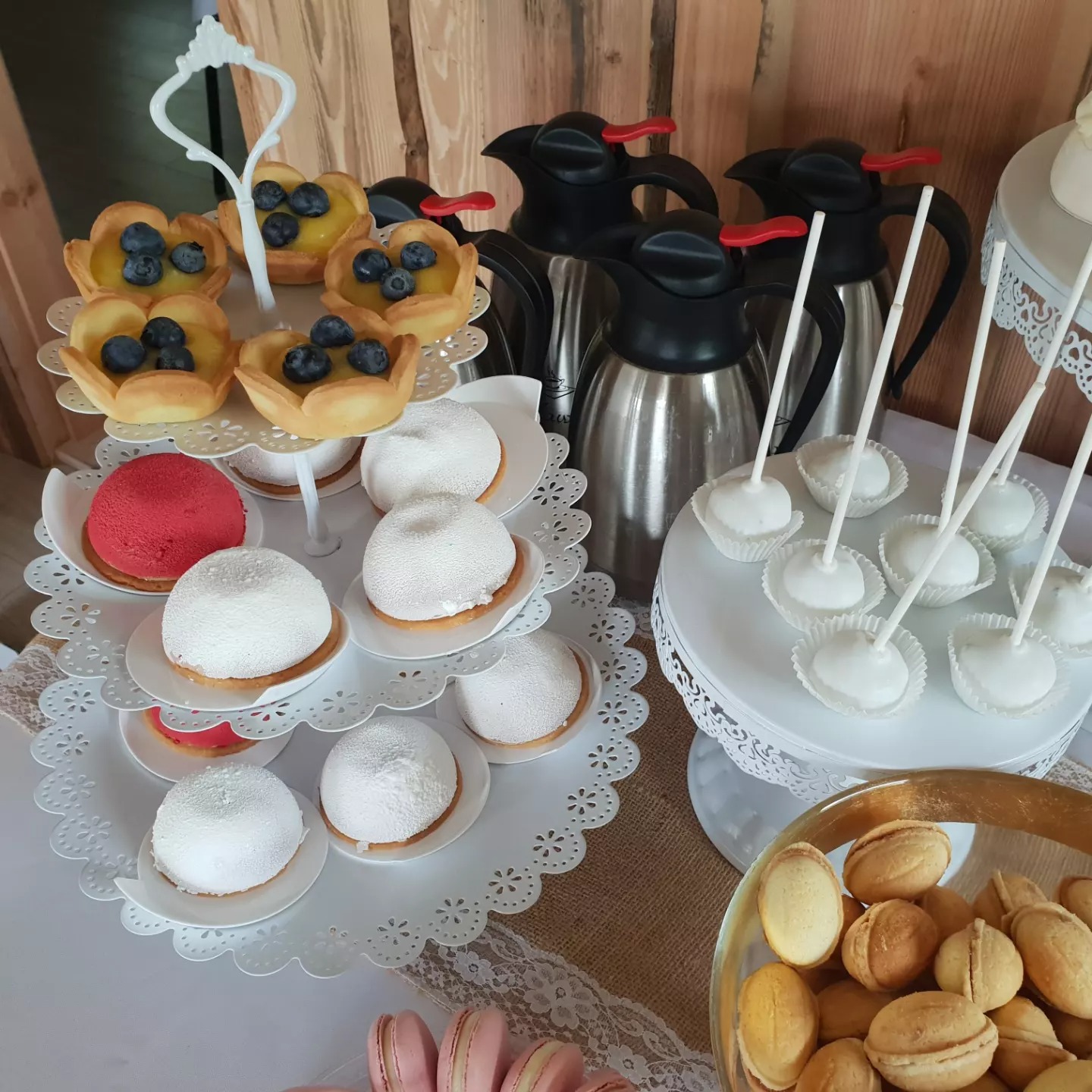 Różnorodność deserów na stole: tartaletki z borówkami, białe kuliste ciastka, cake pops, orzechy w miseczce, różowe makaroniki, całość uzupełniona termosami.