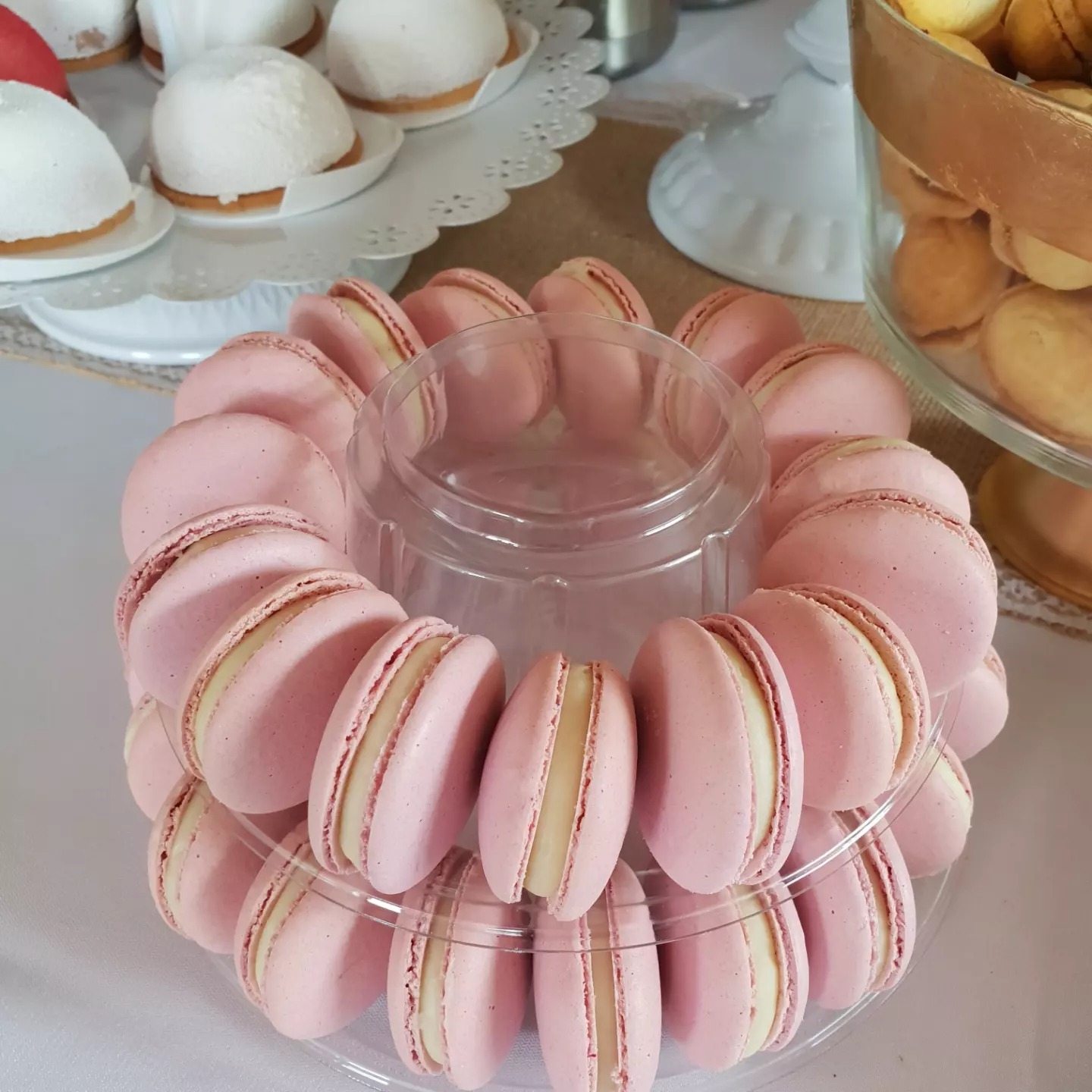 Różowe makaroniki z kremowym nadzieniem ułożone na przezroczystym, dwupoziomowym stojaku, w tle inne desery.