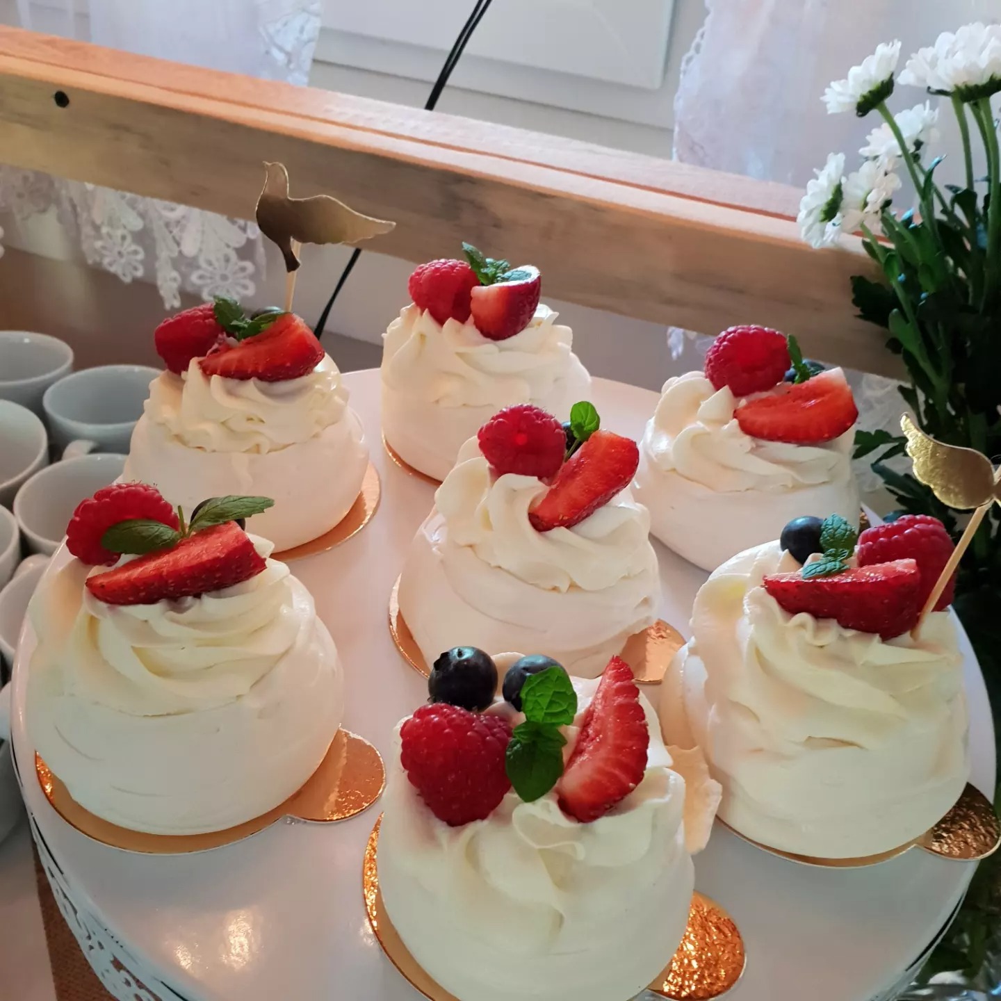 Siedem mini bezowych tortów Pavlova z bitą śmietaną, truskawkami, malinami i borówkami, udekorowanych listkami mięty i złotymi ozdobami na białym paterze, w tle białe kwiaty i filiżanki.