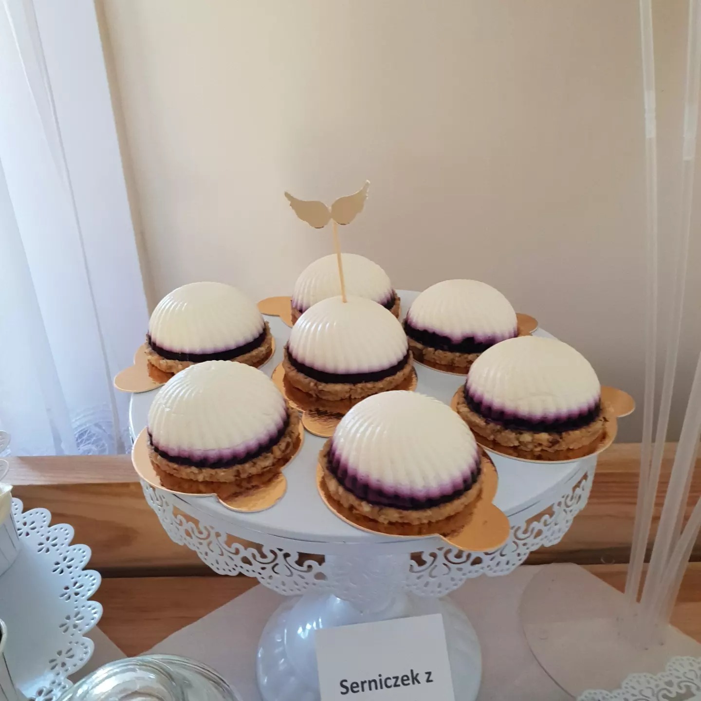 Siedem mini serniczków z galaretką jagodową i białą polewą, ustawionych na białym, ażurowym stojaku; jeden z serniczków ozdobiony złotymi skrzydłami.