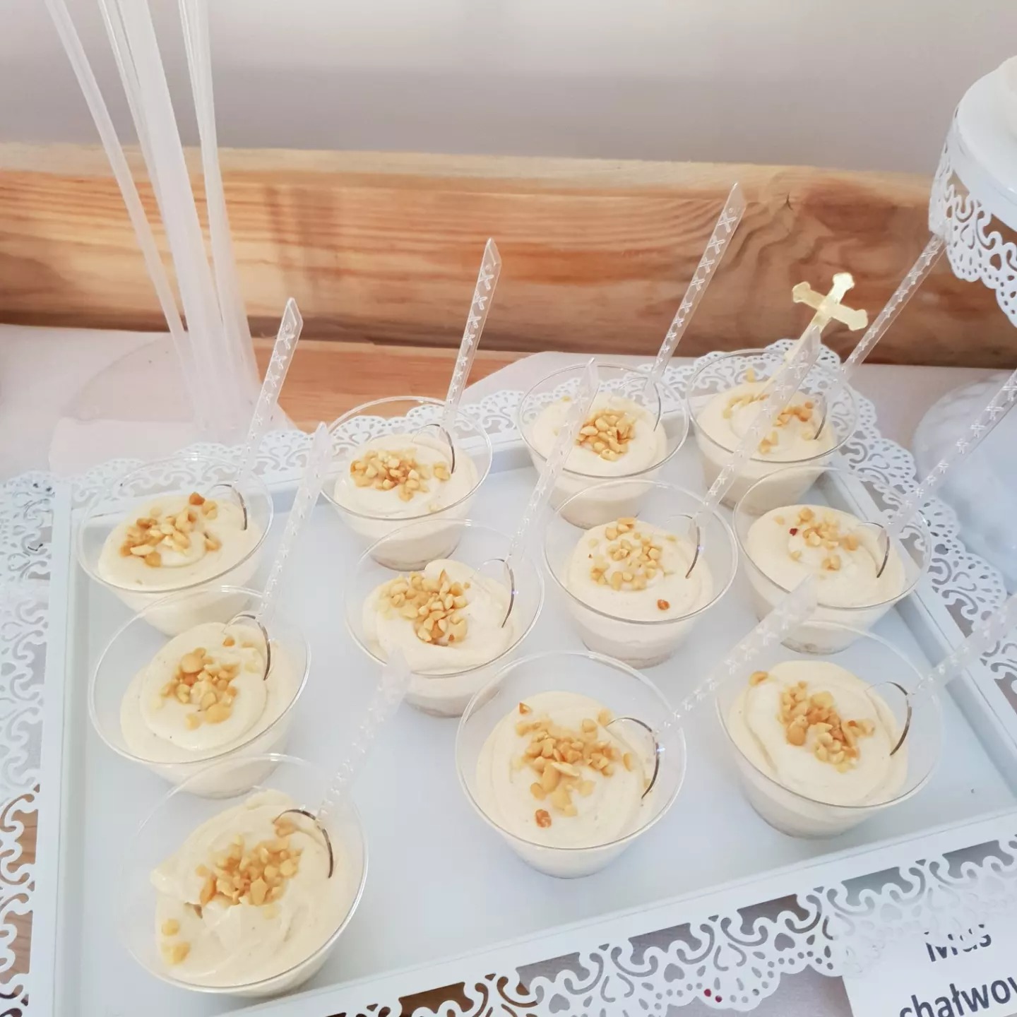 Deser w przezroczystych pucharkach z kremem i posypką orzechową, podany na białej tacy dekorowanej ażurowym wzorem, z przezroczystymi łyżeczkami, jeden z deserów z krzyżykiem.