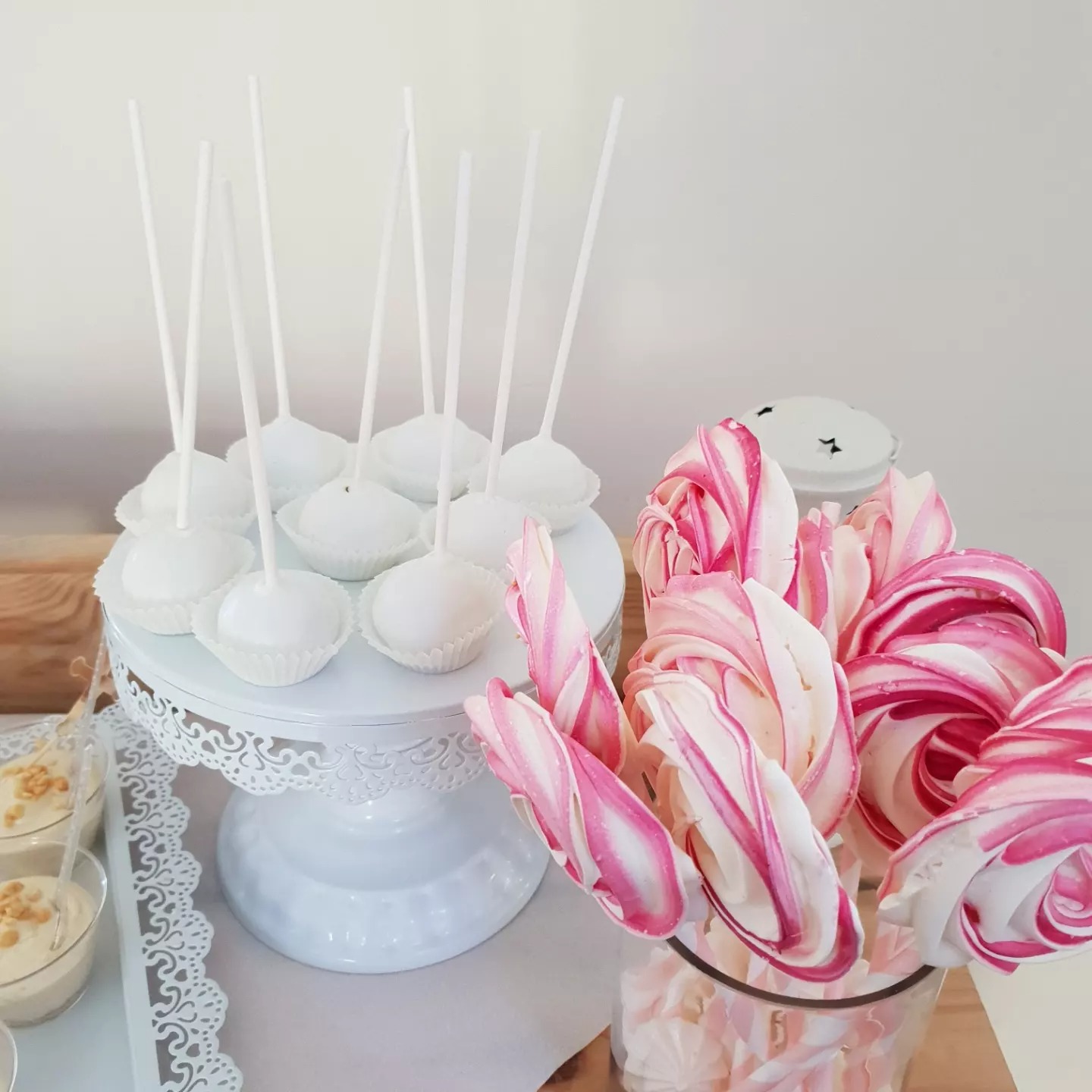 Białe cake pops na ażurowym stojaku, lody deserowe w małych szklankach i lizaki w różowo-białe paski w przezroczystym wazonie.