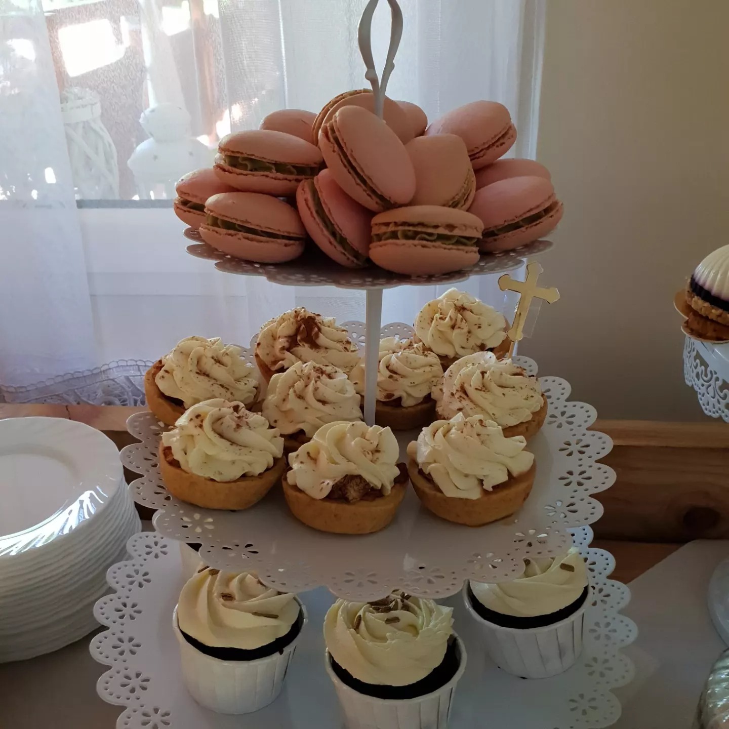 Dwupoziomowa patera z różowymi makaronikami na górze i babeczkami z kremem na dole, obok stos białych talerzy i deser z galaretką, w tle okno z białymi firankami.