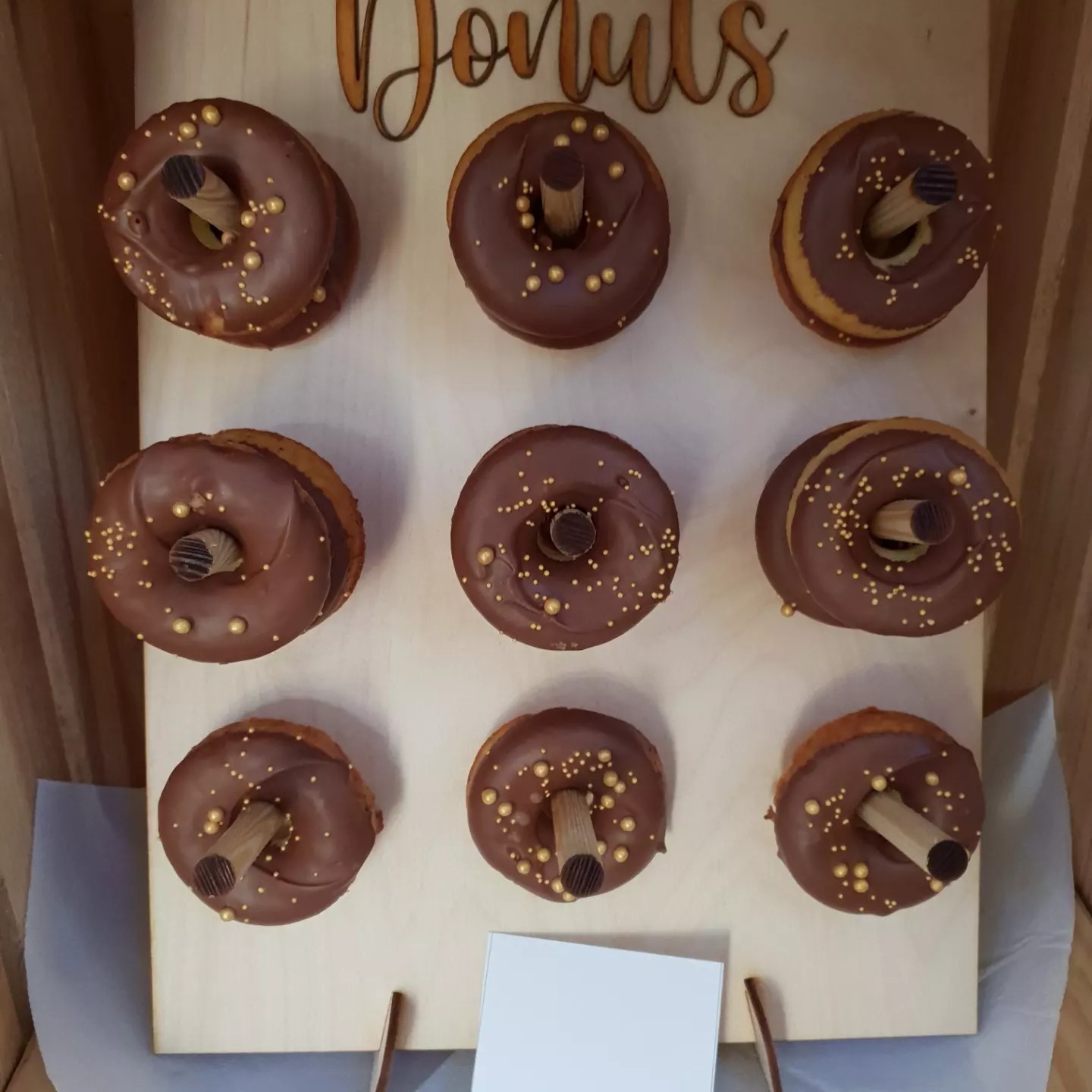Dekoracyjny drewniany stojak na dziewięć pączków z czekoladową polewą i złotymi kuleczkami, każdy pączek umieszczony na drewnianym kołku, z napisem 'Donuts' u góry.