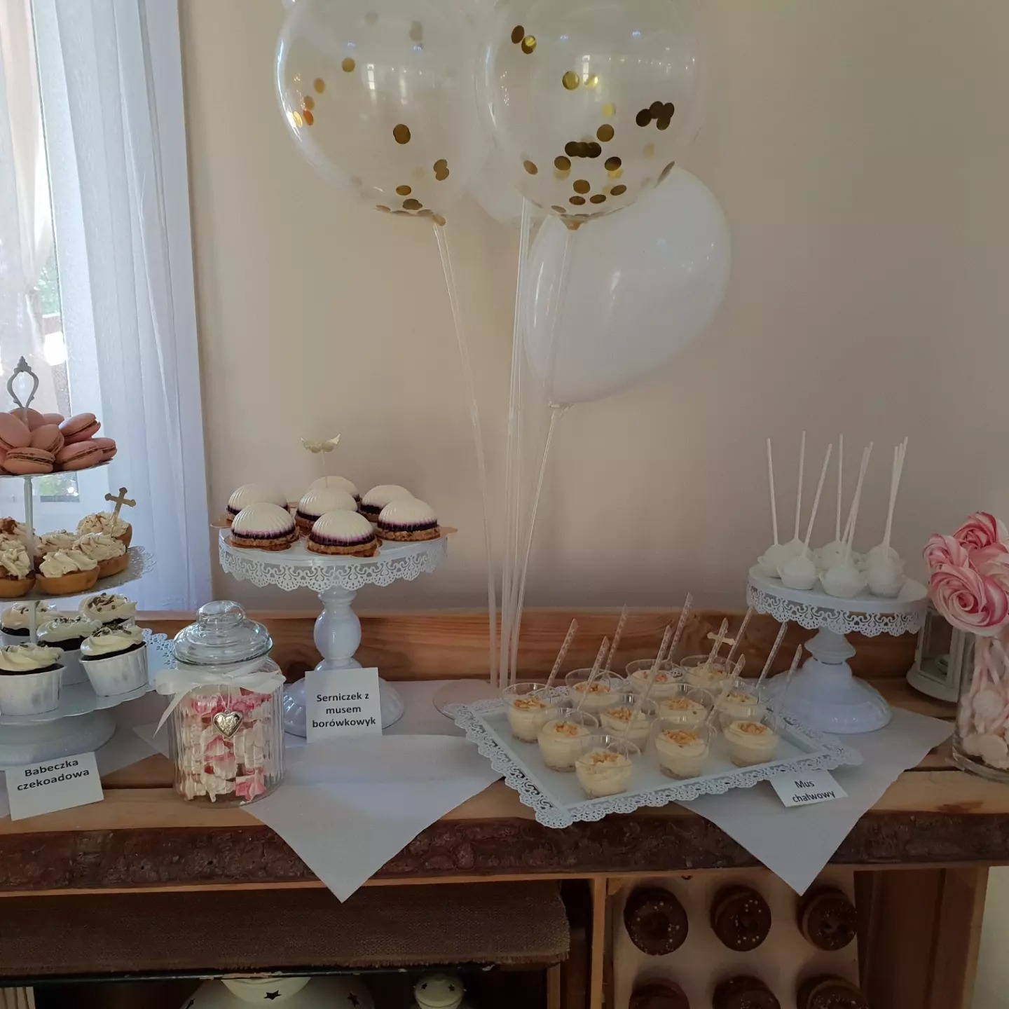 Dekoracja słodkiego stołu z babeczkami czekoladowymi, serniczkiem z musem borówkowym, musem chałwowym, lizakami i białymi balonami z konfetti na drewnianym stole.