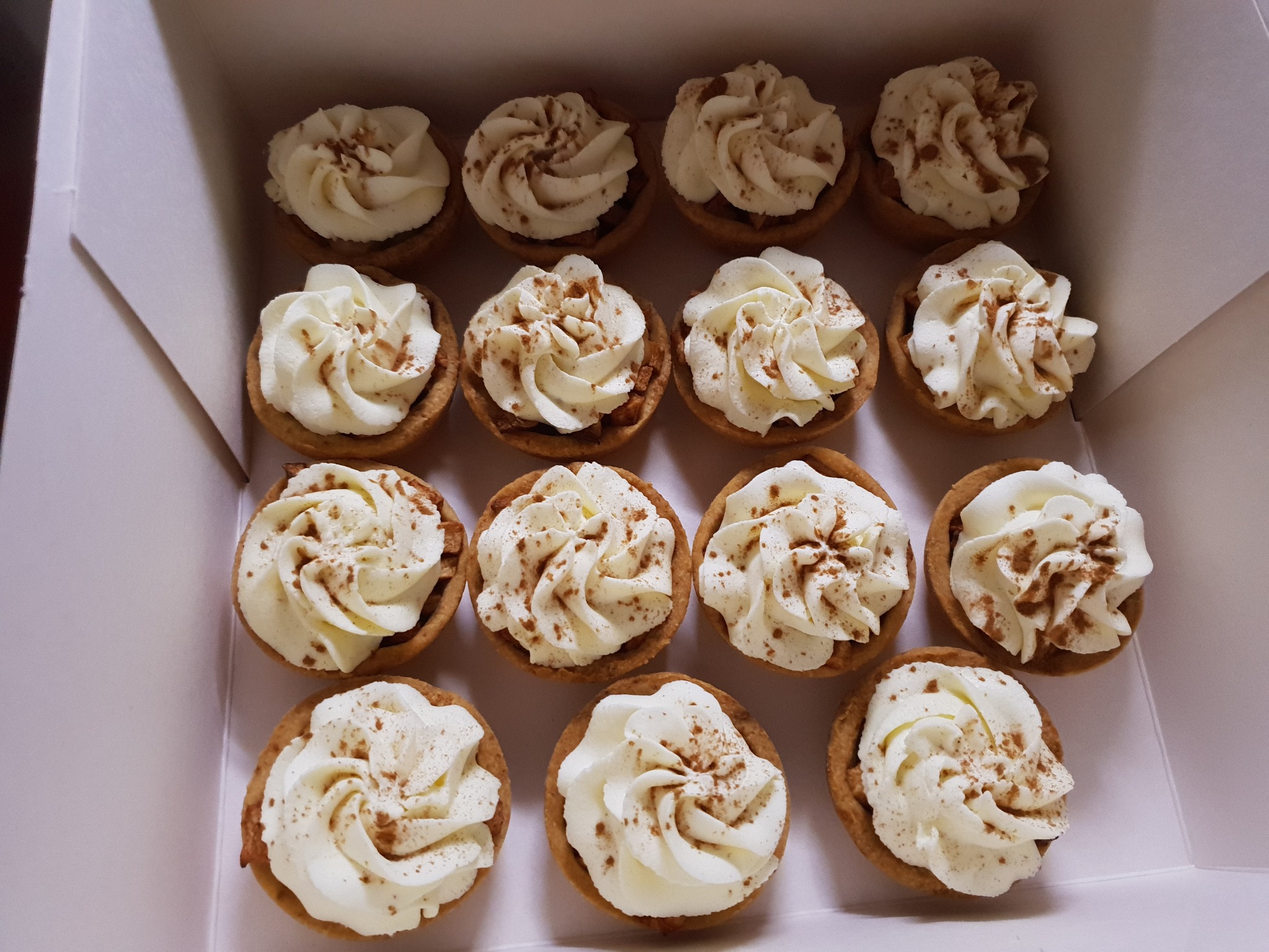 Kartonowe pudełko wypełnione szesnastoma mini tartaletkami z kremem śmietankowym posypanymi cynamonem.