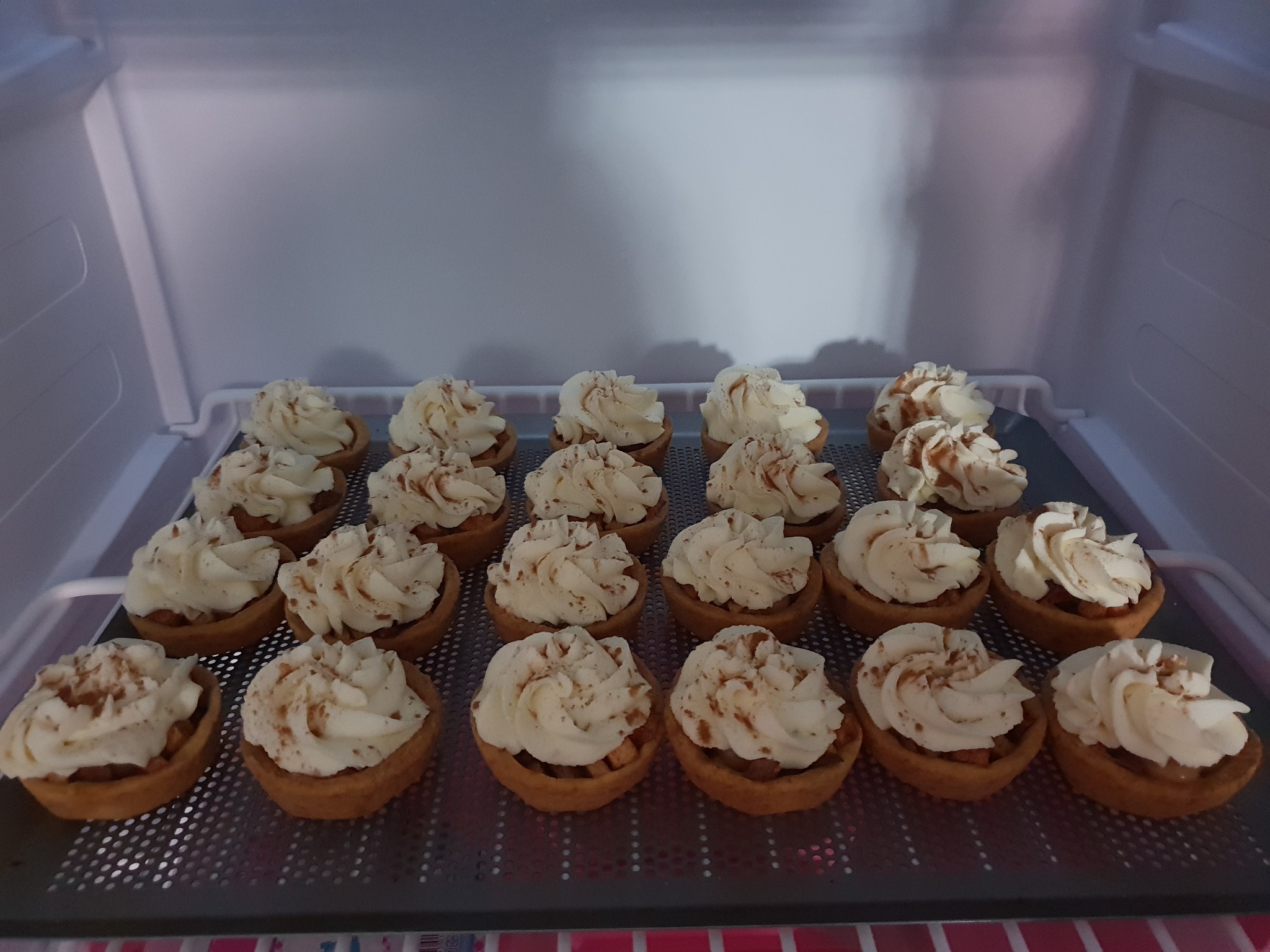 Tarty z jabłkami i bitą śmietaną, posypane cynamonem, ułożone na metalowej tacy w chłodnym oświetleniu lodówki.