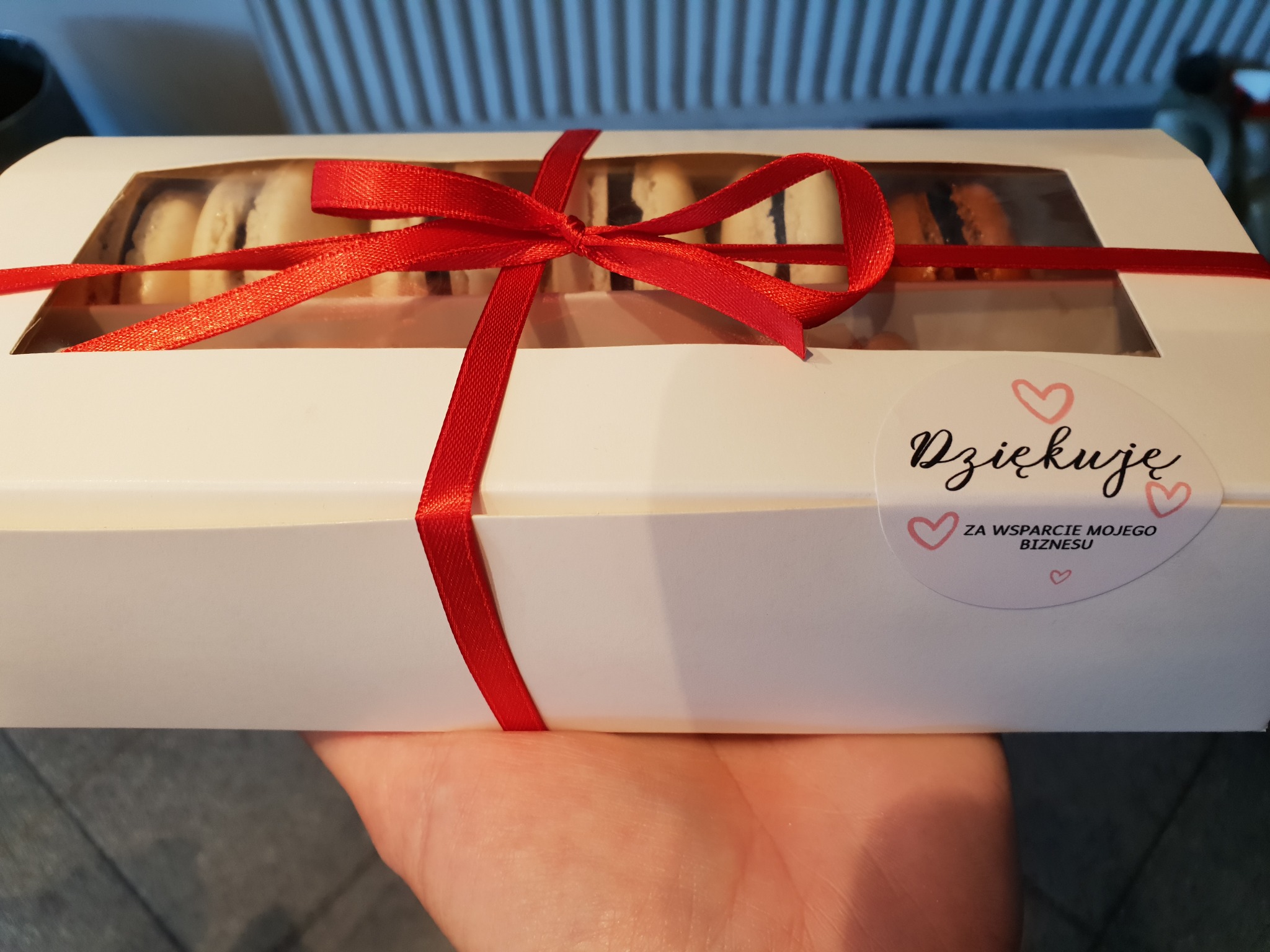 Pudełko białych i brązowych makaroników przewiązane czerwoną wstążką, trzymane w dłoni, z naklejką 'Dziękuję za wsparcie mojego biznesu'.