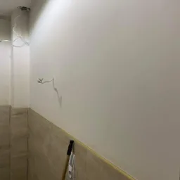 Świeżo wygładzona ściana w trakcie remontu łazienki z wystającymi przewodami elektrycznymi i fragmentem drewnianej rączki narzędzia opartą o ścianę z brązowymi płytkami.