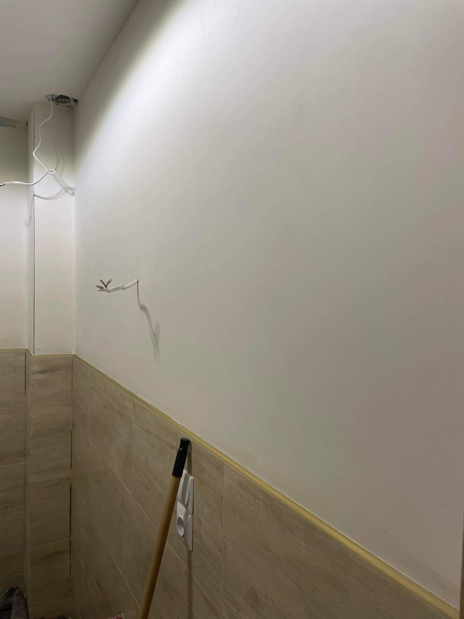 Świeżo wygładzona ściana w trakcie remontu łazienki z wystającymi przewodami elektrycznymi i fragmentem drewnianej rączki narzędzia opartą o ścianę z brązowymi płytkami.