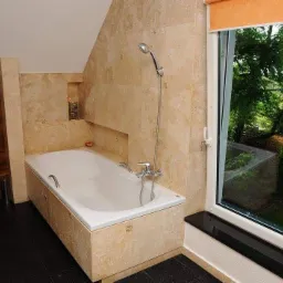 Jasna łazienka na poddaszu z wanną obudowaną trawertynem, toaleta podwieszana na tle ściany z drewnianą okładziną, okno z widokiem na zieleń.