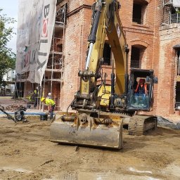 RENOVA GARDEN GROUP SPÓŁKA Z OGRANICZONĄ ODPOWIEDZIALNOŚCIĄ - Żółta koparka na placu budowy z ceglanym budynkiem w tle, pracownicy w odblaskowych kamizelkach, widoczna zagęszczarka wibracyjna i przygotowany teren.