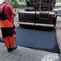 RENOVA GARDEN GROUP SPÓŁKA Z OGRANICZONĄ ODPOWIEDZIALNOŚCIĄ - Pracownicy w odblaskowych kombinezonach obsługują maszynę do rozkładania asfaltu na chodniku z kostki brukowej, widoczne narzędzia i samochody w tle.