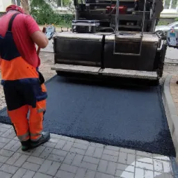 Pracownicy w odblaskowych kombinezonach obsługują maszynę do rozkładania asfaltu na chodniku z kostki brukowej, widoczne narzędzia i samochody w tle.