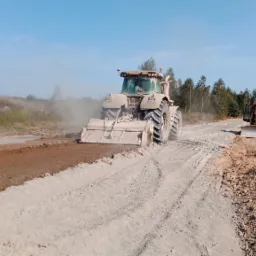 Ciężki traktor z glebogryzarką stabilizuje grunt na budowie drogi, w tle widoczny spychacz i drzewa.