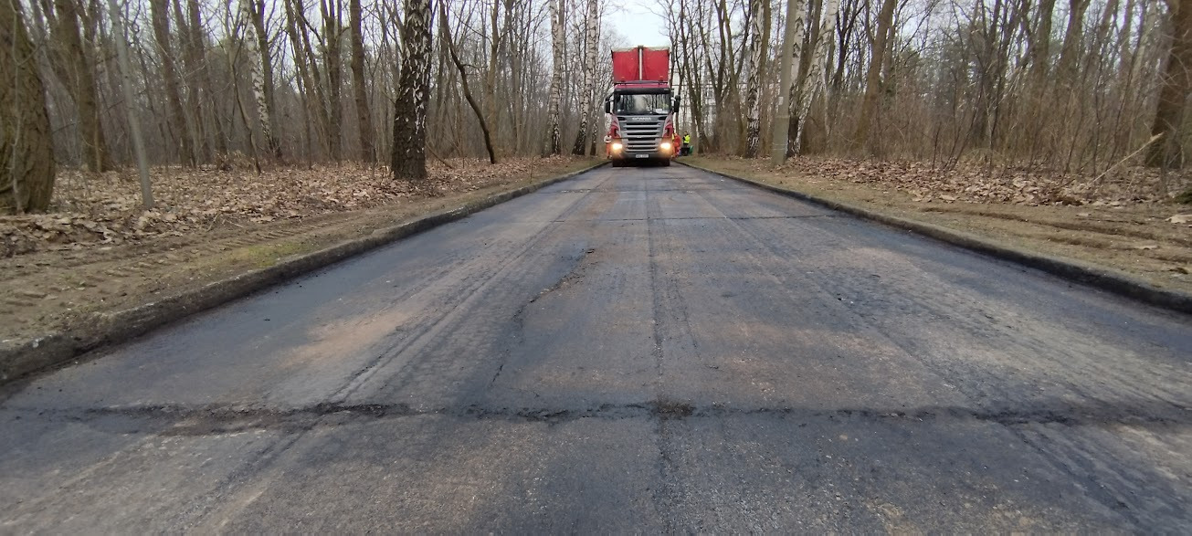Świeżo wyasfaltowana droga w lesie, z widocznymi śladami opon i czerwonym samochodem ciężarowym w oddali.