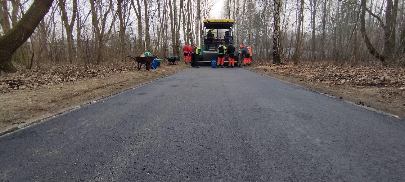 Ekipa robotników w odblaskowych ubraniach przy maszynie Vogele podczas układania świeżego asfaltu na drodze leśnej, w tle drzewa bez liści i wózki transportowe.