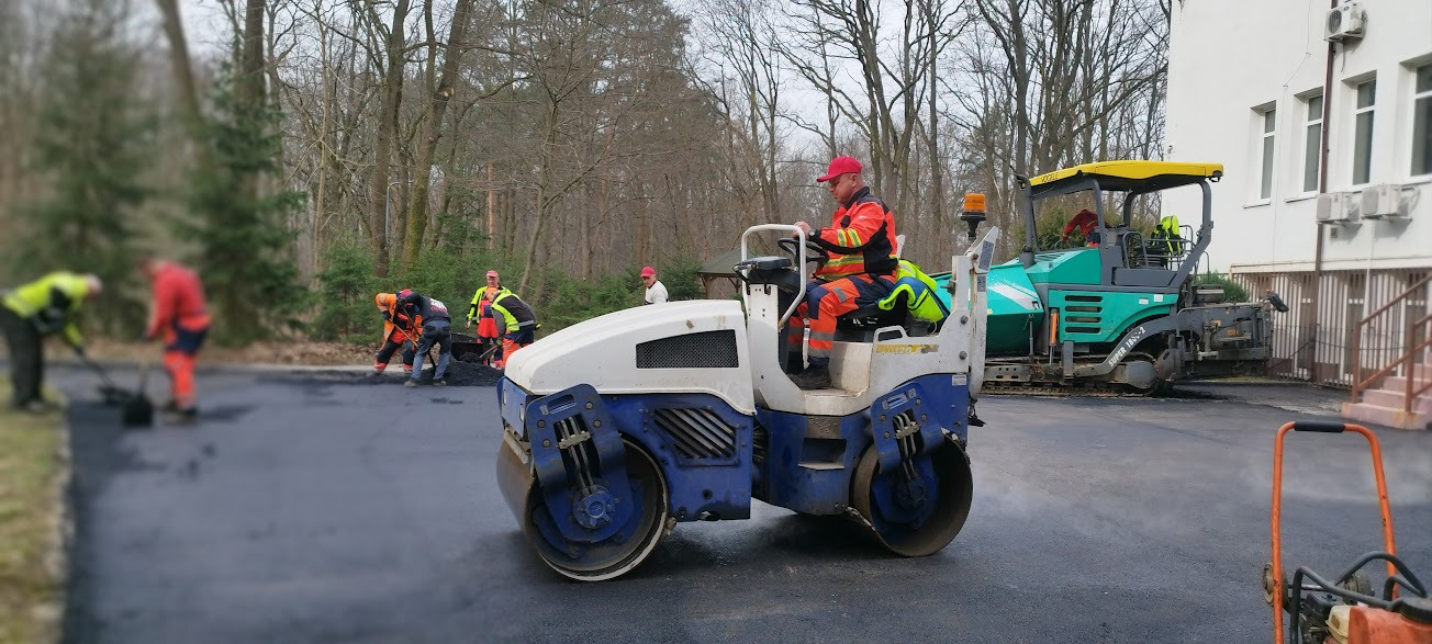 Operator w czerwonej czapce i kombinezonie obsługuje walec drogowego na świeżo wylanym asfalcie. W tle ekipa robotników wyrównuje nawierzchnię, a obok stoi rozściełacz asfaltu.