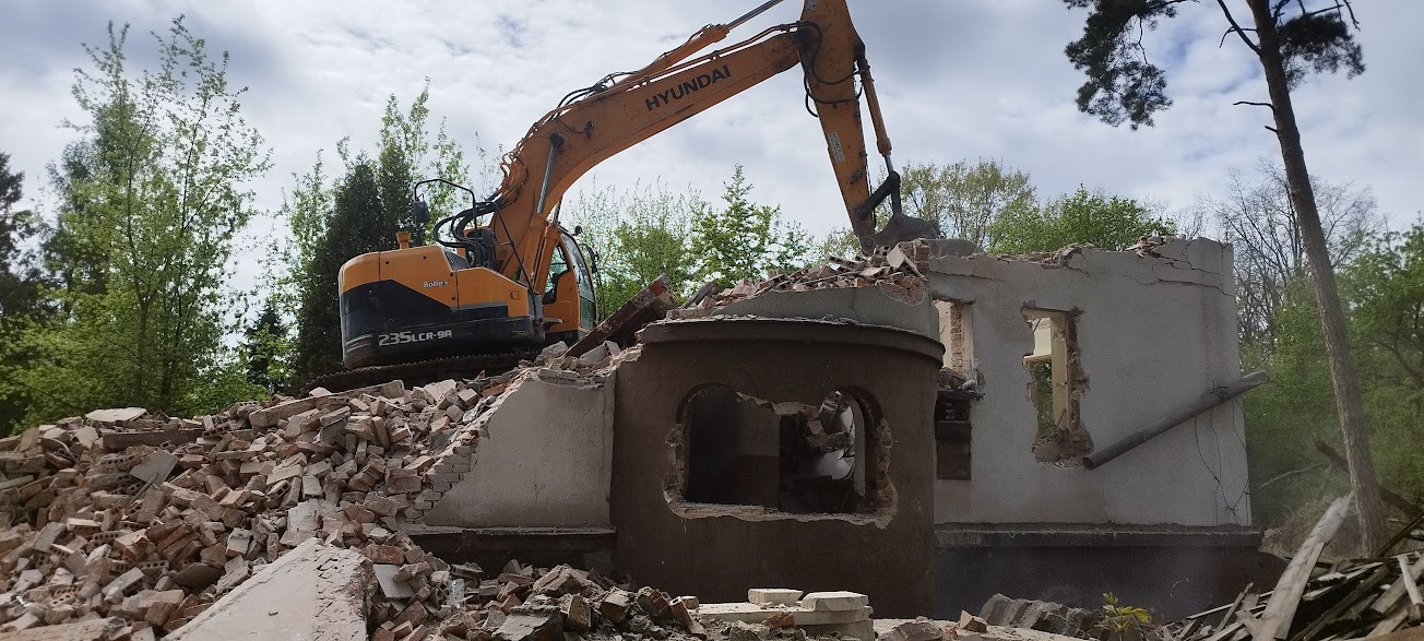 Pomarańczowa koparka Hyundai 235LCR-9A na gruzach budynku z cegły, widok z boku na tle drzew i pochmurnego nieba.