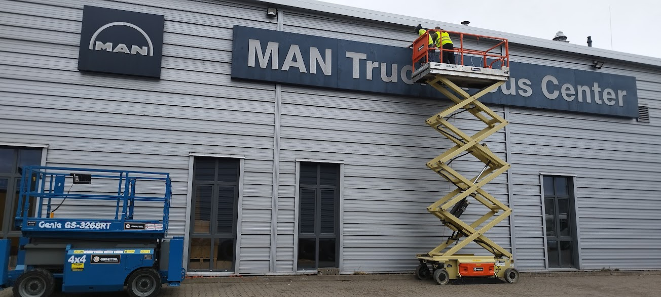 Pracownicy na podnośniku koszowym montują logo MAN Truck & Bus Center na elewacji budynku z szarej blachy trapezowej, w tle błękitny podnośnik Genie GS-3268RT.