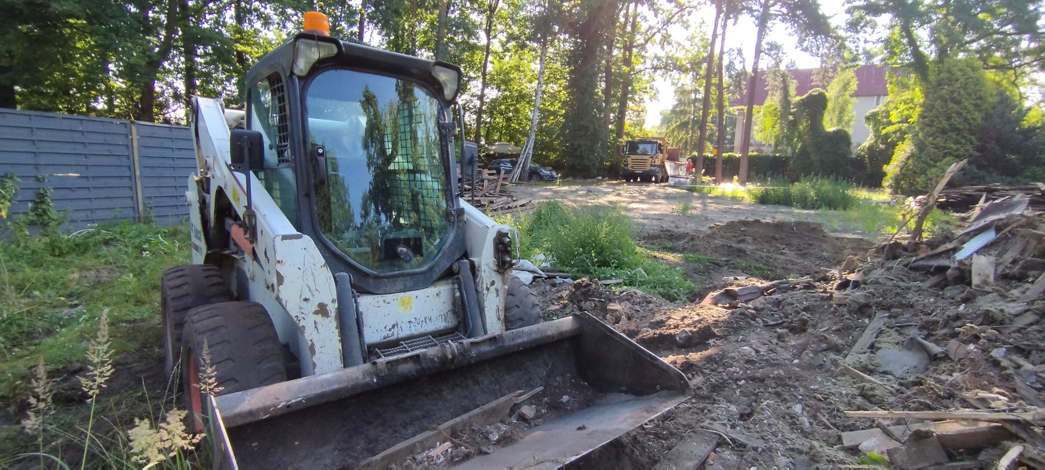 Biała ładowarka Bobcat na placu budowy, widoczna sterta gruzu i wywrotka w tle, otoczenie drzewa i ogrodzenie.