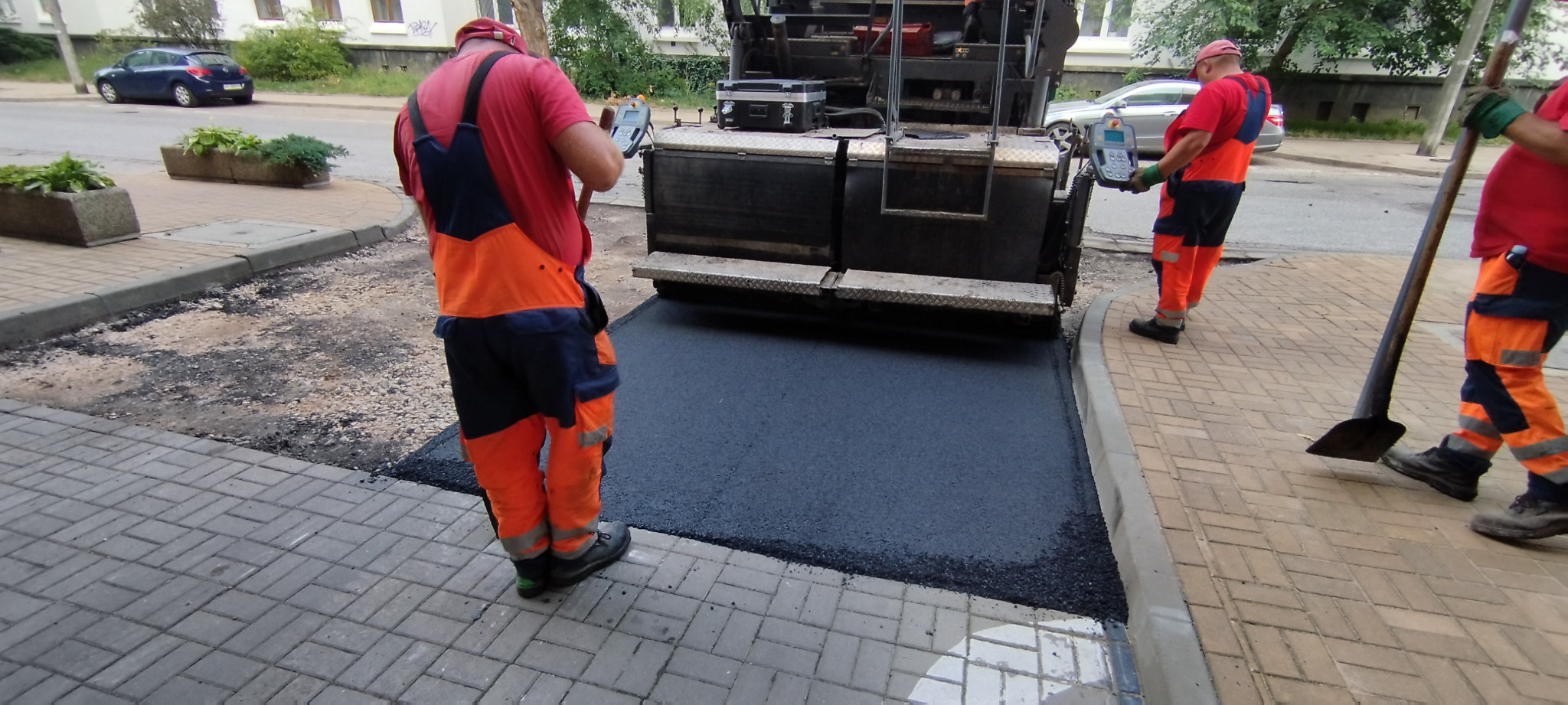 Pracownicy w odblaskowych kombinezonach obsługują maszynę do rozkładania asfaltu na chodniku z kostki brukowej, widoczne narzędzia i samochody w tle.