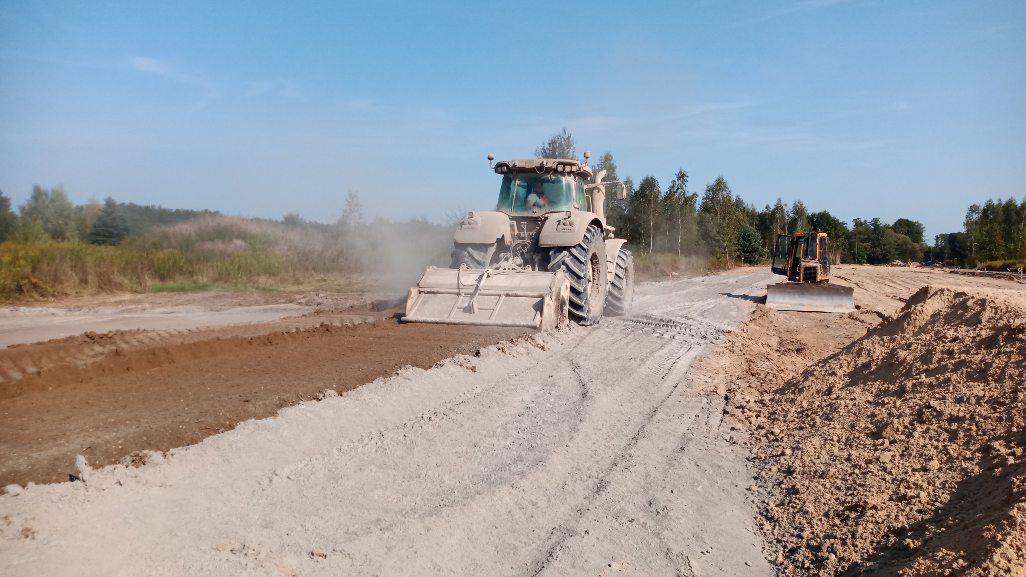 Ciężki traktor z glebogryzarką stabilizuje grunt na budowie drogi, w tle widoczny spychacz i drzewa.
