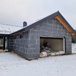 Dom jednorodzinny w trakcie prac ociepleniowych, ściany pokryte grafitowym styropianem z widocznymi kołkami, zimowy krajobraz w tle, śnieg na dachu i wokół budynku.