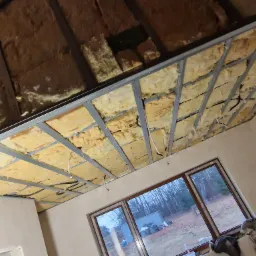 Sufit poddasza w trakcie izolacji wełną mineralną, widoczna metalowa konstrukcja nośna, fragment instalacji elektrycznej i okno z widokiem na zewnątrz.