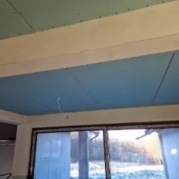 Montaż niebieskich płyt karton gips na suficie w pomieszczeniu z oknem, widoczne ślady po wkrętach i wystający kabel.