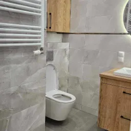 Wykończona łazienka w odcieniach szarości i drewna, z wiszącą toaletą, grzejnikiem drabinkowym i szafką pod umywalką z drewnianym frontem.