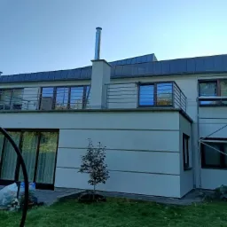 Dwupiętrowy dom z otynkowaną elewacją w odcieniach bieli, z balkonem na pierwszym piętrze i metalowym kominem, na tle błękitnego nieba, z widocznym fragmentem rusztowania.