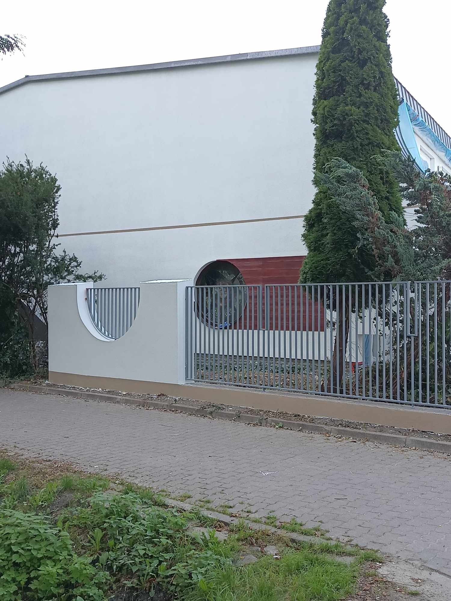 Biała elewacja budynku z nietypowym okrągłym oknem, widoczna zza metalowego ogrodzenia i fragmentu murku z półokrągłym wycięciem, obok rosną drzewa.