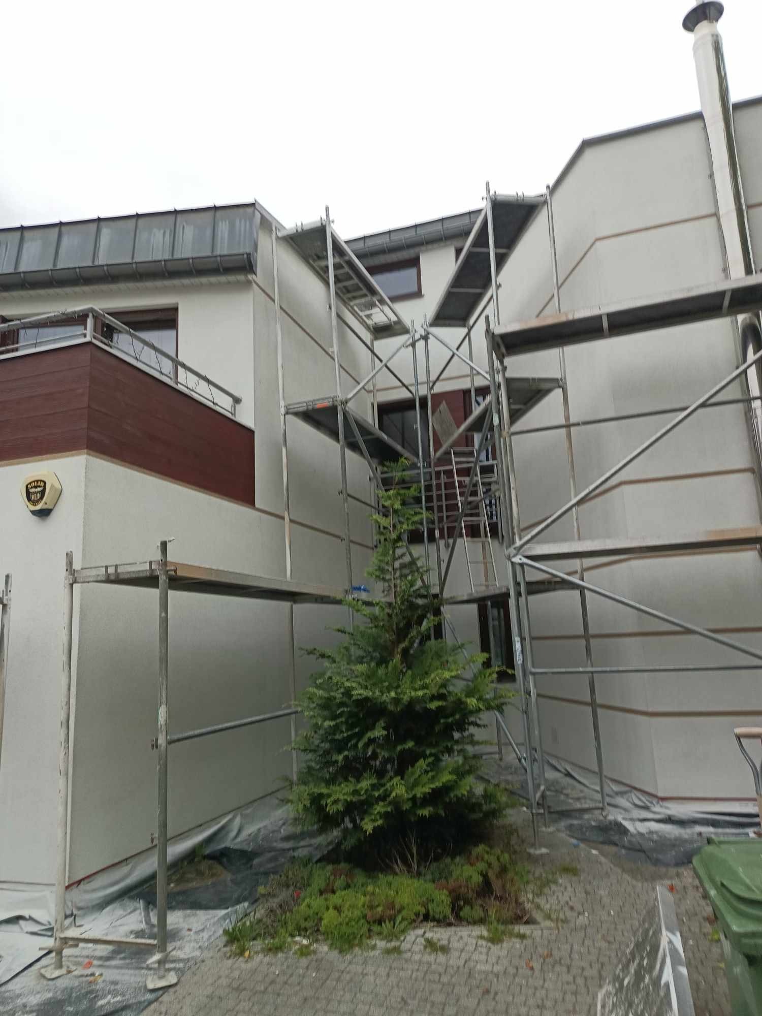 Dom jednorodzinny w trakcie prac elewacyjnych, otoczony rusztowaniem i zabezpieczony folią, z widocznym fragmentem zielonego trawnika i małym drzewkiem