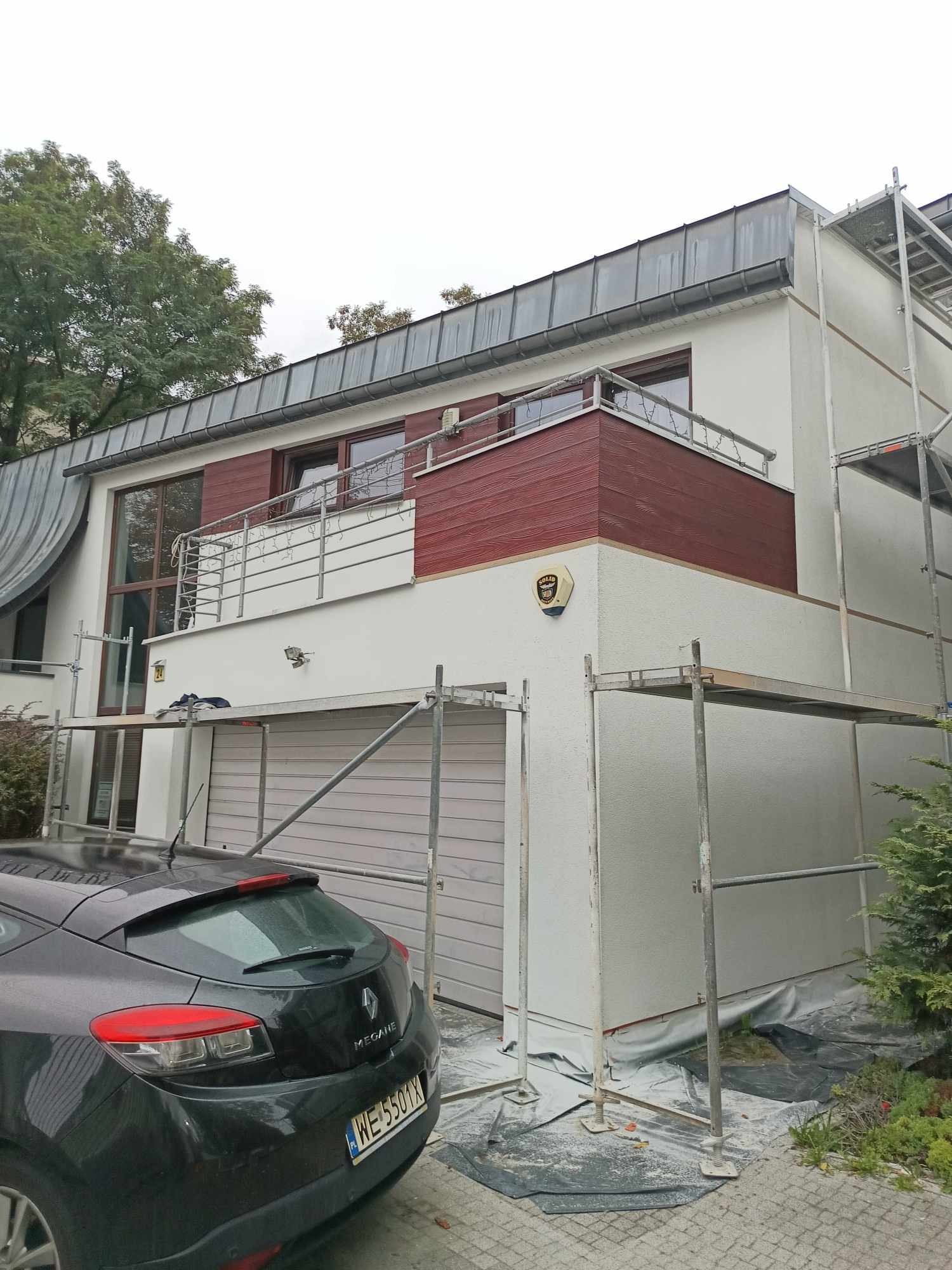 Dom dwukondygnacyjny w trakcie prac elewacyjnych, rusztowania wokół budynku, częściowo zakryte okna, widoczny samochód osobowy na podjeździe.