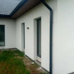 Nowoczesny dom jednorodzinny z jasną elewacją, czarnymi oknami i rynnami, widok z boku na fragment budynku z trawnikiem w pierwszym planie.