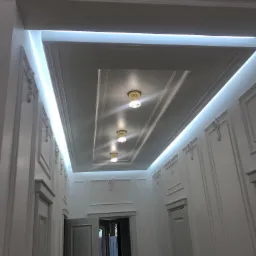 Białe, eleganckie wnętrze z dekoracyjnymi sztukateriami na ścianach i podświetlanym sufitem kasetonowym z trzema kryształowymi lampami, widok z perspektywy korytarza z otwartymi drzwiami.