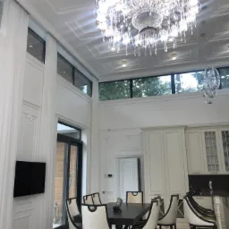 Eleganckie wnętrze jadalni z wysokim sufitem, ozdobną sztukaterią, kryształowym żyrandolem i widokiem na zieleń za oknem.