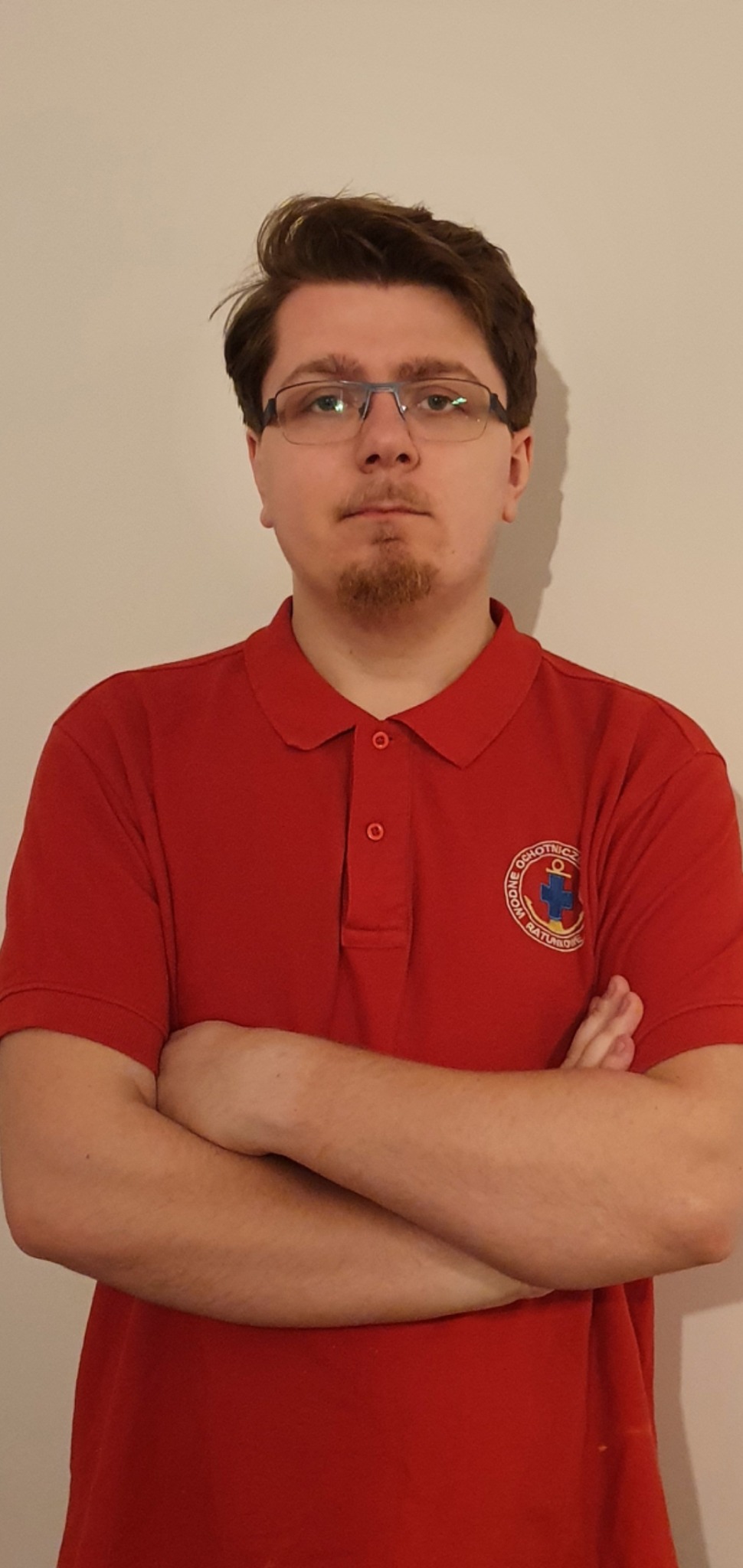 Portret mężczyzny w czerwonej koszulce polo z logo WOPR, skrzyżowane ręce, neutralne tło.