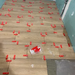 Układanie płytek podłogowych imitujących drewno z użyciem czerwonych klipsów poziomujących i białych krzyżyków dystansowych. Widoczna warstwa kleju. Ściany przygotowane pod glazurę.