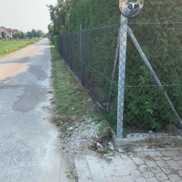 Michał Stawarski - Metalowa siatka ogrodzeniowa z okrągłym lustrem drogowym na słupku, odbijającym fragment ulicy i samochód. Tło: zielone tuje.