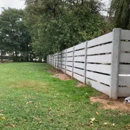 Szary, betonowy płot lamelowy z szerokimi przerwami między deskami, biegnący wzdłuż trawnika z drzewami w tle, widziany z perspektywy żabiej z dołu.